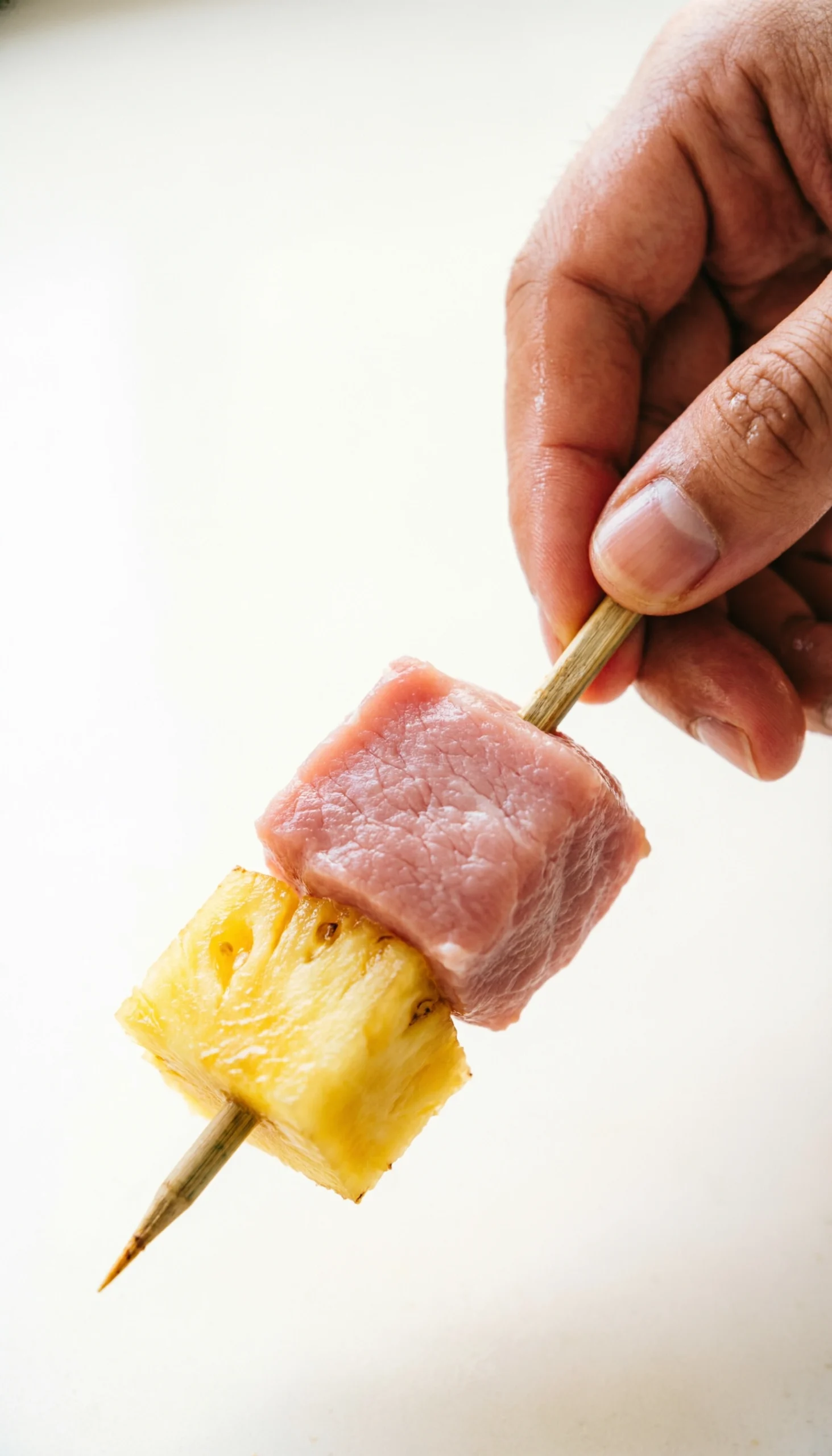 A Hand Threads A Juicy Pork Cube And Fresh Pineapple Onto A Bamboo Skewer, Sharply Focused In Bright Daylight On A Clean Prep Area.