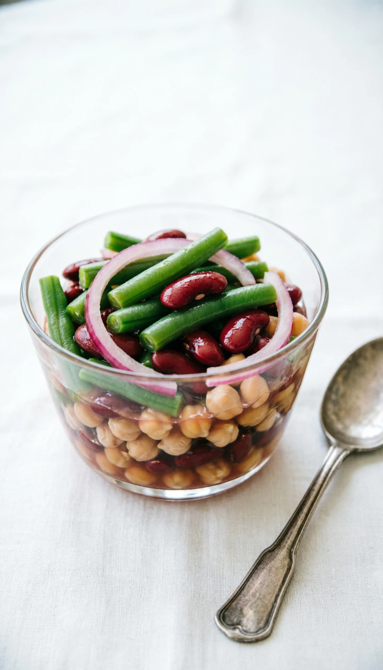 Classic layered three-bean salad with green beans, kidney beans, and chickpeas in a glass trifle bowl on white linen.