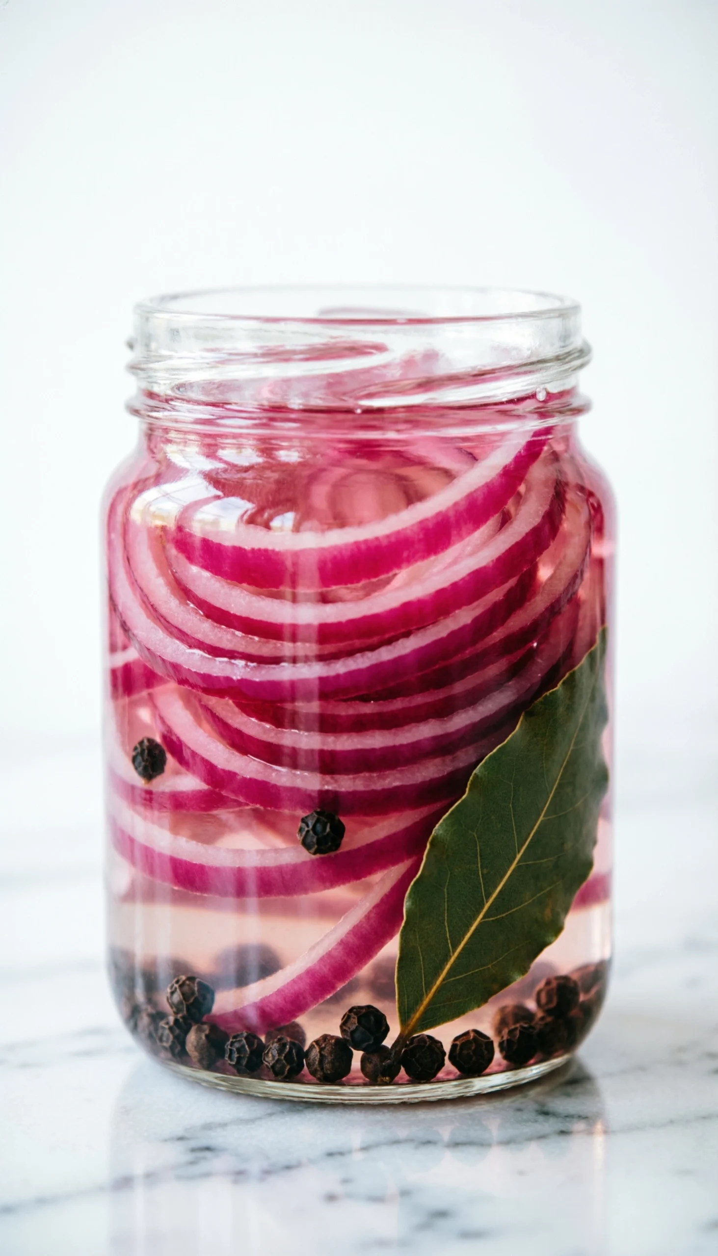 Sliced pink pickled red onions in a clear glass jar on a white marble countertop with peppercorns and bay leaves.