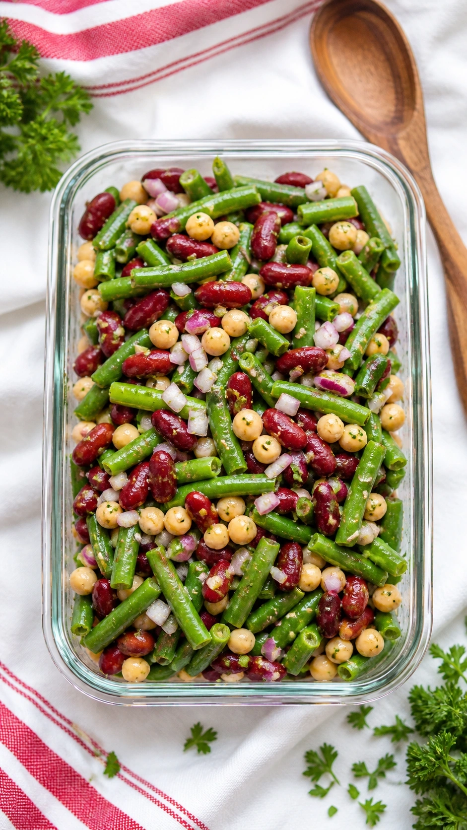 Vibrant three bean salad with green snap beans, kidney beans, garbanzo beans, and red onion in a glass container on a picnic blanket.